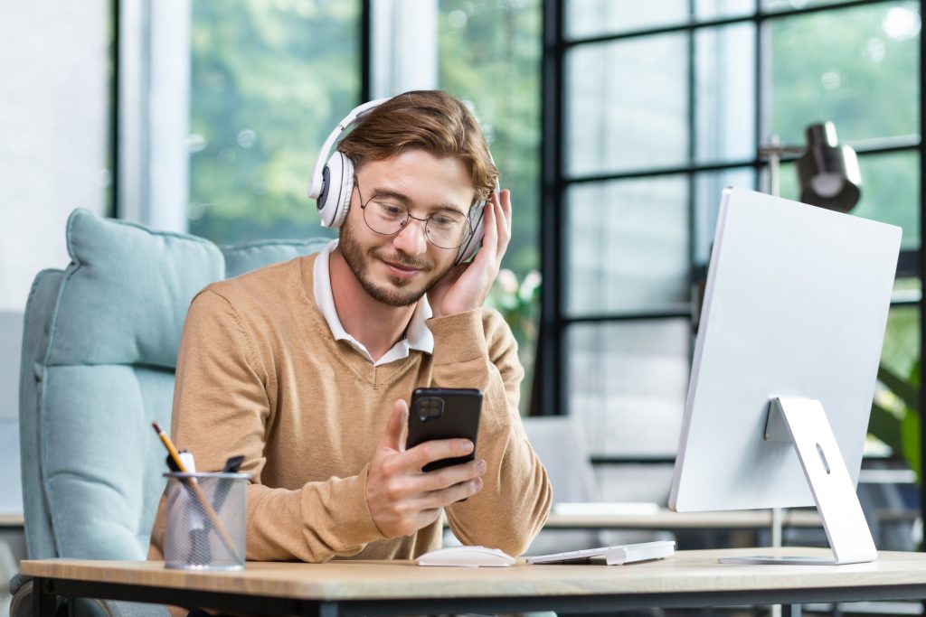 A young man at the workplace in headphones holds a phone in his hand, sits at a table in the office, listens to music, a podcast, an audio book during a break between work, rests